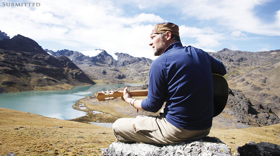Survivorman Les Stroud playing guitar in scenic area with mountains and river in the background.
