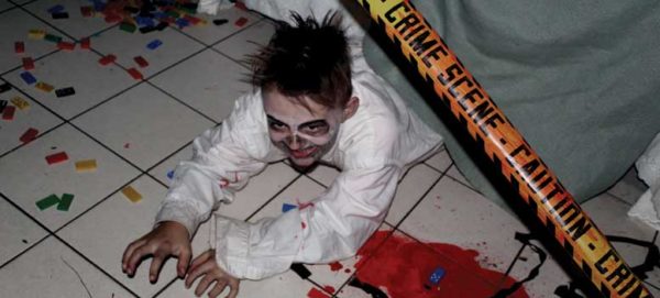 Boy with skeleton facepaint and fake blood on face lays on haunted house floor, half covered by a tarp and surrounded by legos.