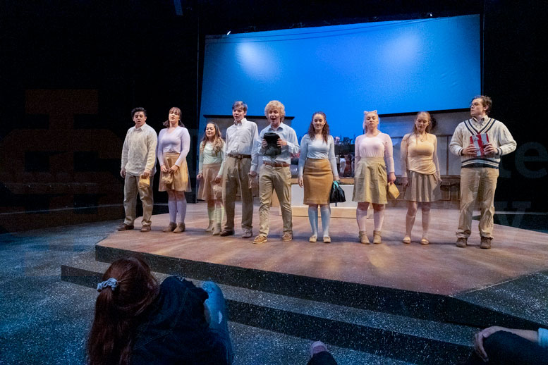 Cast members stand on the edge of the stage, facing the audience