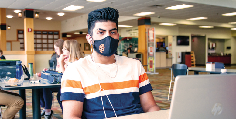 Image of Diego Cortez, sitting at a table in the PSUB, wearing a black mask with an ISU insignia
