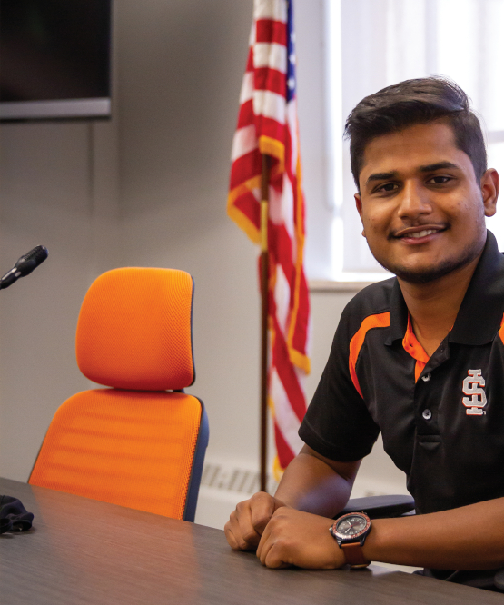 Aayush Jha, ASISU President sits at a table with an empty orange chair next to him and an AMerican flag in the background.