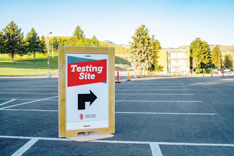 A wooden sandwich board reading "Testing Site" with an arrow pointing to the right stands in the empty parking lot used for COVID testing.