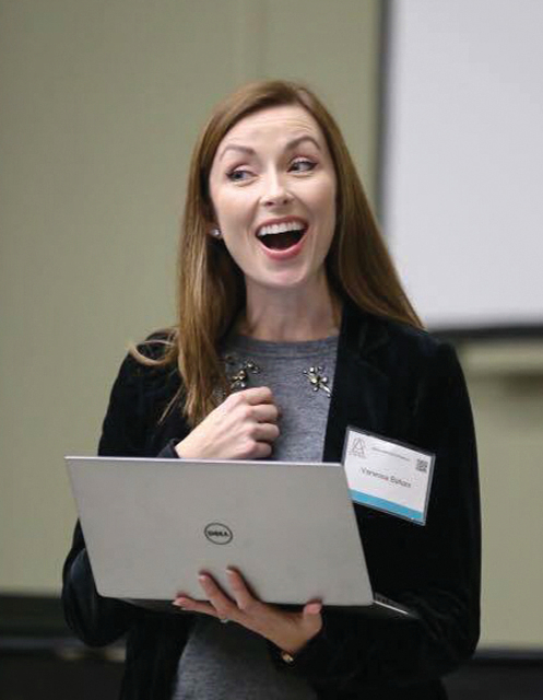 Vanessa Ballam holds a laptop in her left hand while her right hand is held in a loose fist over her chest. Her mouth is open as if she is speaking or singing.