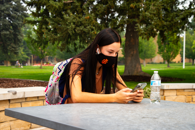 Girls wearing face mask sits at table outside on quad while looking at her phone.