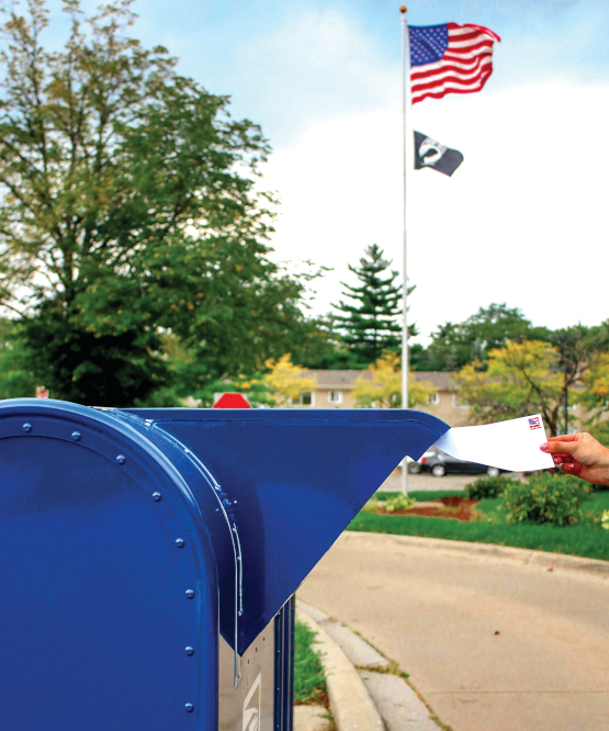 Photo of person dropping letter into blue mail box. ALl that can be seen of the person is their hand. A flag pole with the US flag is visible in the background.