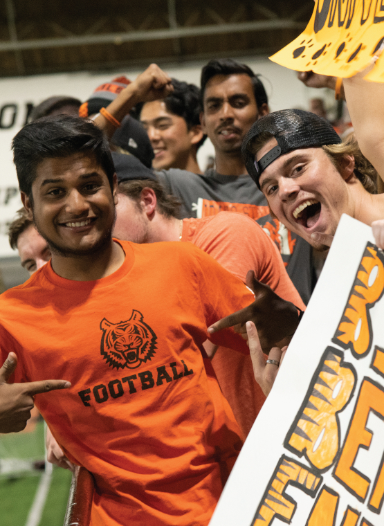 Diego Cortez, wearing and orange Bengal shirt, points his fingers at the logo. Another student leans into the shot, smiling. Other students stand behind, out of focus.