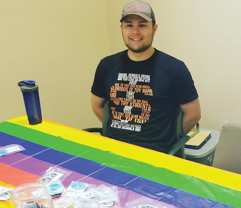 Student sits behind table. He is wearing a hat and a t-shirt with an ISU logo on it. He is smiling and has his arms tucked behind him. On the table, there's a rainbow flag tablecoth and various flyers and handouts scattered. There is also a blue water bottle sitting on the table.