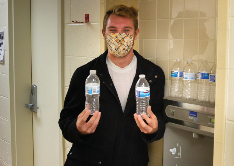 Zach Jewkes stands next to drinking fountain with a mask on. He is holding an empty plastic disposable water bottle in each hand, with four sitting on top of the drinking fountain behind him.