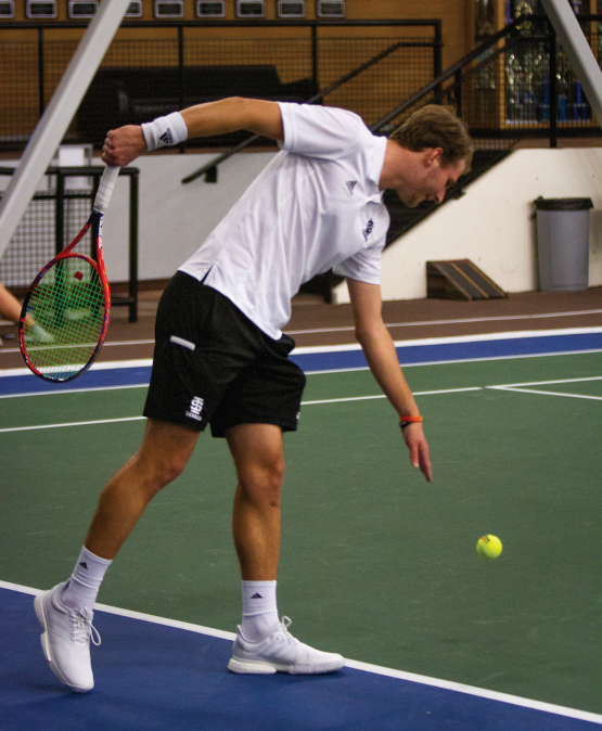 Gary Rendek, on a tennis court and holding a racket, leans over to bounce a tennis ball before he serves.