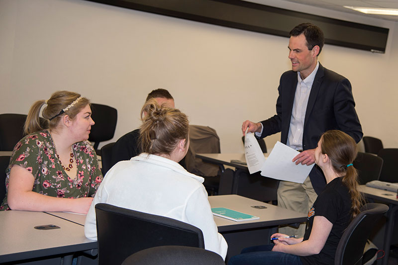 Alex Bolinger stands with papers in his hands, talking with three female students and one male student. They are seated around a table and only their backs and profiles can be seen.