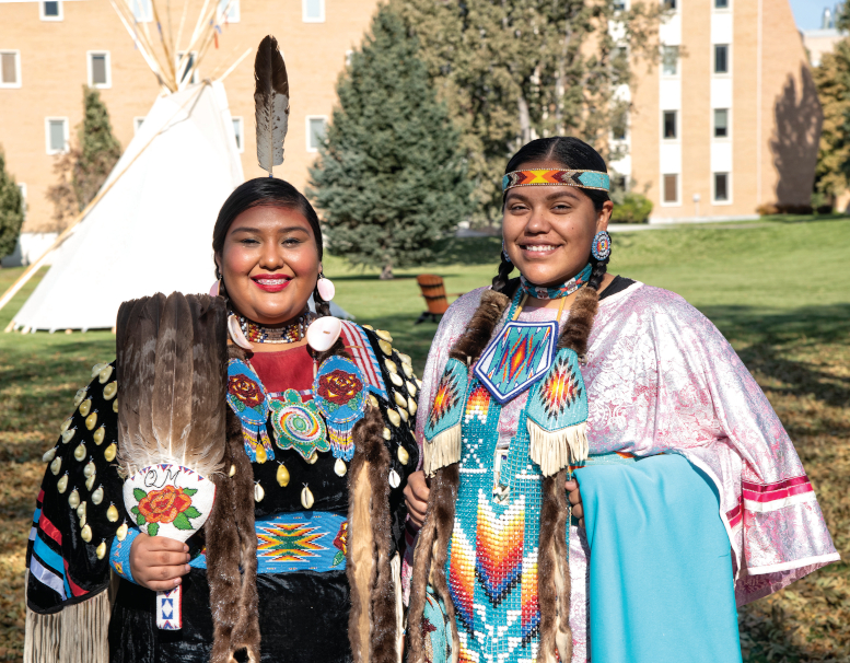 Two Native American women pose smiling, dressed in traditional garb.