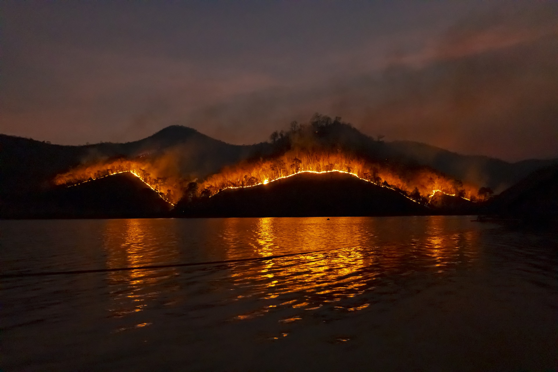 Photo at night, shot is taken looking at hills/mountains across a body of water. The landscape is burning.