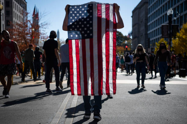 Person stands on street with crowd of people behind them. They are holding up an American flag so it covers their whole body except their feet.