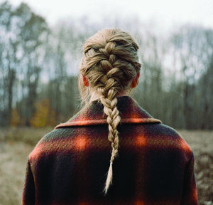 Photo of Swift from behind, her hair in a braid down her back. She is wearing a plaid coat and looking out into a wooded area.