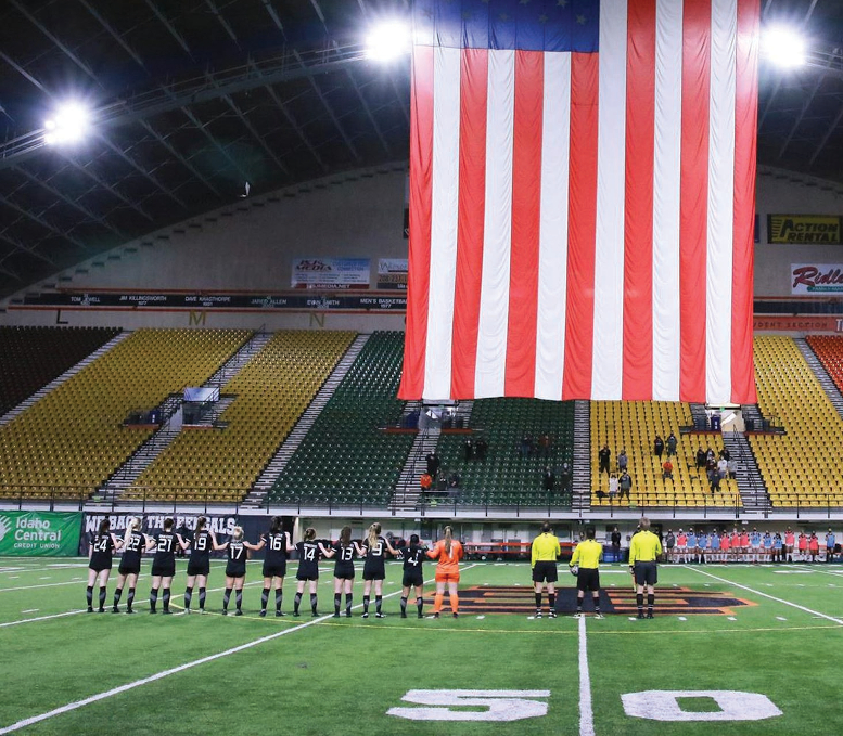 Soccer players stand on field with American flag overhead