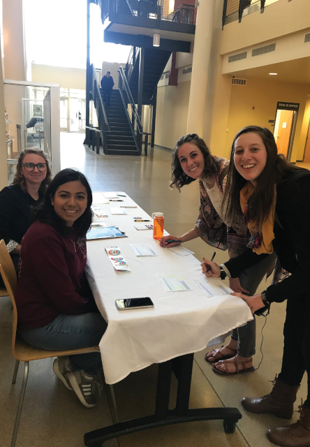 Two women sit at a table, turned towards camera, smiling. Two other women lean over table, signing something and smiling