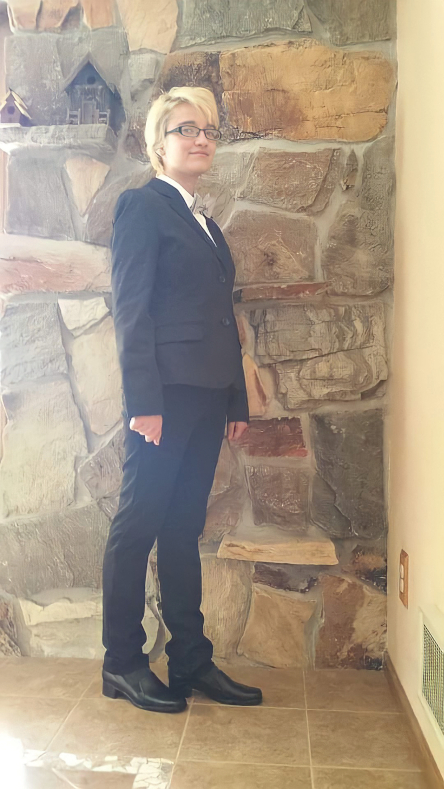 Lesley Brey in a black suit with short blond hair and glasses stands with her right shoulder angled towards the camera