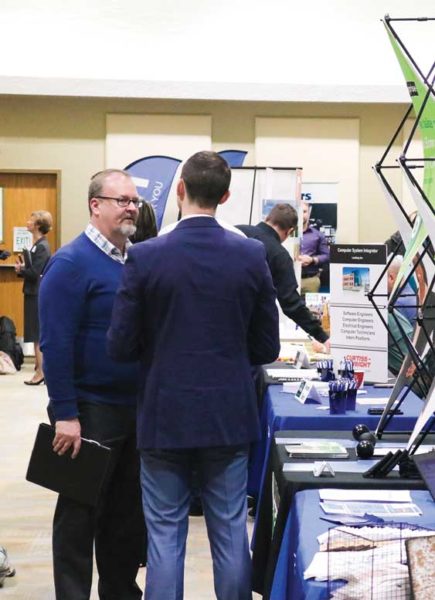 Two men speak to each other while standing in front of a table at the career fair.