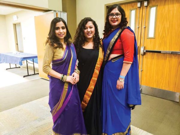 Three women stand together, dressed in saris