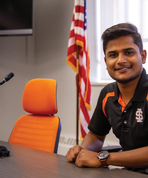 Aayush Jha, ASISU President sits at a table with an empty orange chair next to him and an AMerican flag in the background.
