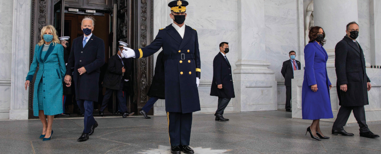 Man in uniform stands with arm gesturing to his right. On his right side President Joe Biden and First Lady Jill Bide walk. On his other side, Vice President Kamala Harris and Second Gentleman Doug Emhoff